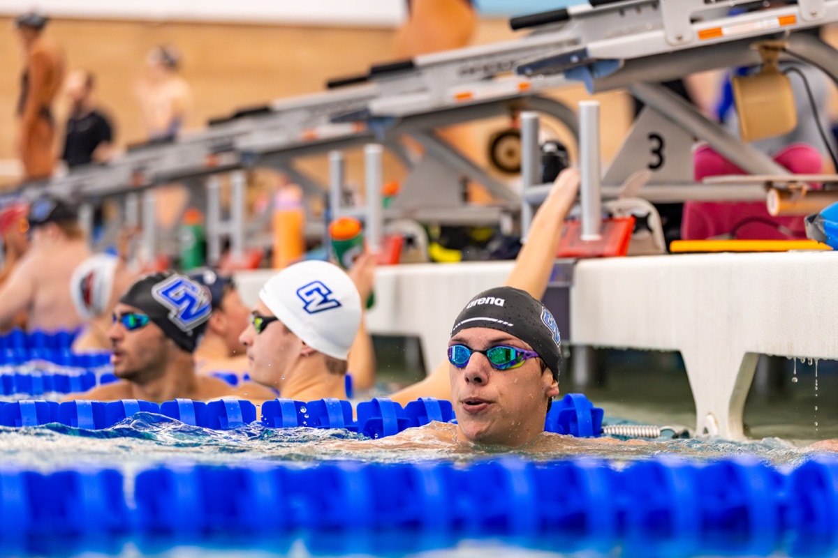 Tom in the pool in a black arena cap and mirrored goggles, hand on the lane rope